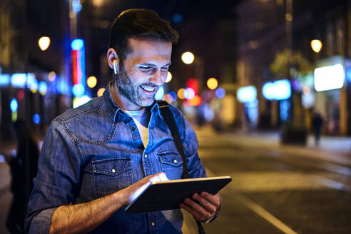Lächelnder Mann mit drahtlosen Kopfhörern, der in der nächtlichen Stadt ein digitales Tablet benutzt - BSZF01118