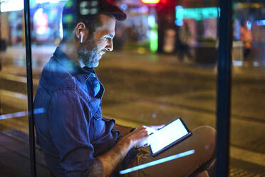 Mann mit Kopfhörern, der nachts an einem Bahnhof sitzt und sein digitales Tablet benutzt - BSZF01119