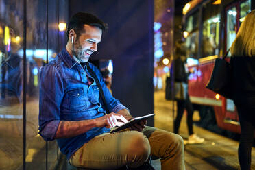 Man with headphones sitting at a station at night using his digital tablet - BSZF01120