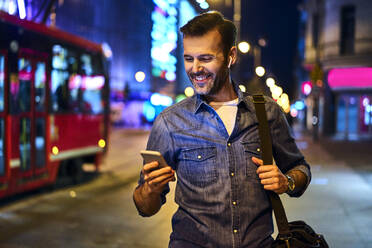 Lächelnder Mann mit drahtlosen Kopfhörern, der sein Smartphone benutzt, während er nachts durch die Stadt läuft - BSZF01121