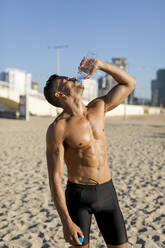 Mann mit nacktem Oberkörper trinkt Wasser nach dem Training am Strand - MAUF02665