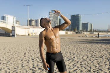 Mann mit nacktem Oberkörper trinkt Wasser nach dem Training am Strand - MAUF02666