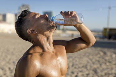 Mann mit nacktem Oberkörper trinkt Wasser nach dem Training am Strand - MAUF02667