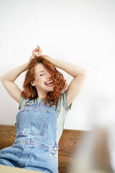 Cheerful redhead woman sitting with arms raised against white wall at home - DMOF00154