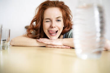 Porträt einer fröhlichen Frau, die zwinkert und die Zunge herausstreckt, während sie sich zu Hause auf den Tisch stützt - DMOF00156