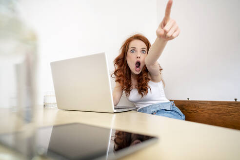 Schockierte Frau, die schreiend auf einen Tisch mit Laptop zeigt und gegen eine weiße Wand im Zimmer sitzt - DMOF00168