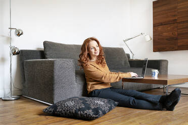 Full length of woman sitting with laptop while looking away in living room at home - DMOF00172