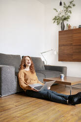 Full length of cheerful woman laughing while sitting with laptop in living room at home - DMOF00173