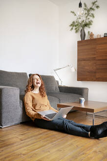Fröhliche Frau lacht in voller Länge, während sie mit einem Laptop im Wohnzimmer zu Hause sitzt - DMOF00173