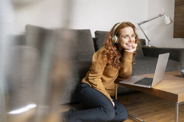 Porträt einer schönen Frau mit Kopfhörern, die mit ihrem Laptop auf dem Couchtisch sitzt - DMOF00176
