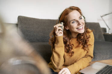 Smiling beautiful woman wearing headphones while sitting by laptop on coffee table at home - DMOF00178