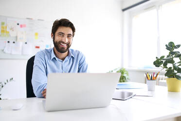 Portrait of a smiling businessman sitting at a desk using a laptop - BSZF01129