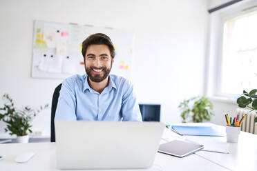 Porträt eines lächelnden Geschäftsmannes, der an einem Schreibtisch sitzt und einen Laptop benutzt - BSZF01130