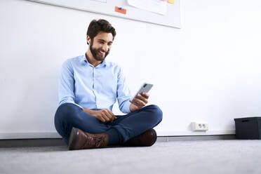 Lächelnder Geschäftsmann, der auf dem Boden sitzt und sein Smartphone benutzt - BSZF01144