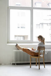 Junge Frau sitzt auf einem Stuhl am Fenster im Büro und macht sich Notizen - JOSF03461