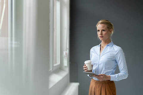 Nachdenkliche junge Geschäftsfrau mit Tablet und Kaffee zum Mitnehmen im Büro, lizenzfreies Stockfoto