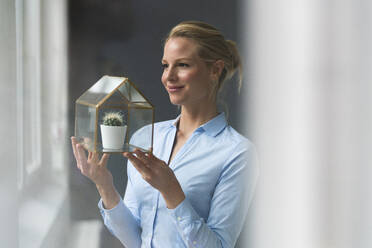 Smiling young businesswoman holding glass house model with cactus inside - JOSF03495