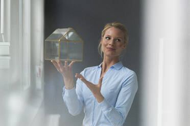 Smiling young businesswoman holding glass house model - JOSF03497