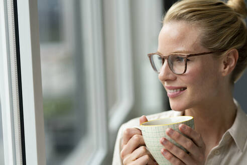 Young businesswoman with cup of coffee looking out of window - JOSF03531