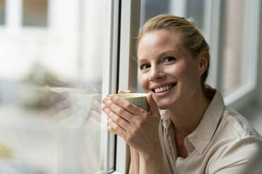 Porträt einer lächelnden jungen Geschäftsfrau mit einer Tasse Kaffee am Fenster - JOSF03533