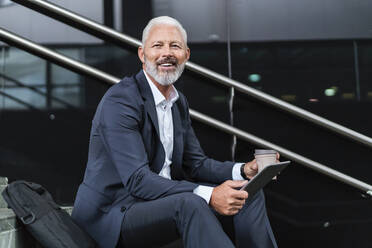 Smiling mature businessman sitting on stairs using tablet - DIGF07414