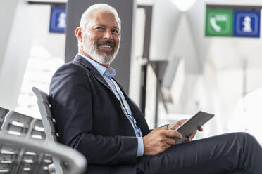 Smiling mature businessman sitting in waiting area with tablet - DIGF07460