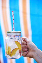 Close-up of Woman's hand holding drink in mason jar filled with ice and slices of peach - BZF00503