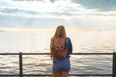 Rear view of woman with backpack standing at railing by sea against sky - TAMF01797