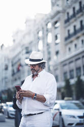 Älterer Mann mit weißer Kleidung und Hut benutzt ein Smartphone in einer Straße in Madrid - OCMF00464