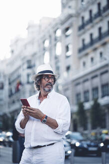 Attraktiver älterer Mann mit weißer Kleidung und Hut, der wegschaut, während er ein Smartphone in einer Straße in Madrid hält - OCMF00465