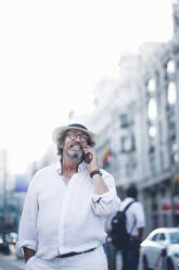 Lächelnder älterer Mann mit weißem Haar und Hut, der wegschaut, während er auf der Straße von Madrid mit einem Smartphone spricht - OCMF00466