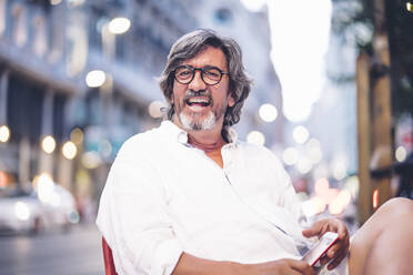 Porträt eines fröhlichen älteren Mannes, der mit seinem Smartphone in einem Straßencafé in Madrid sitzt - OCMF00472