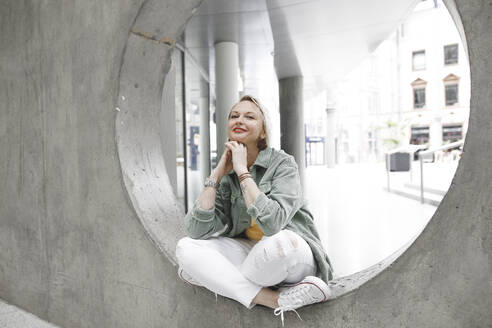 Full Länge Porträt der schönen reifen Frau lächelnd, während auf Beton Kreis in der Wand sitzen - KMKF01008