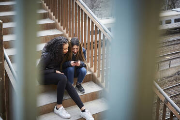 Teenage girl showing mobile phone to female friend while sitting on steps - MASF12835