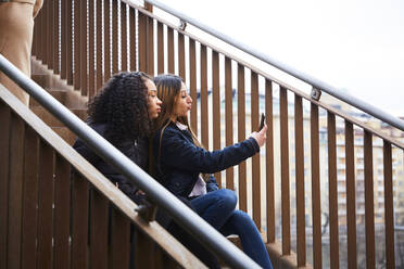 Teenage girl taking selfie with female friend while sitting on staircase - MASF12840