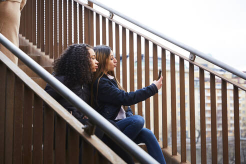 Teenager-Mädchen, das ein Selfie mit einer Freundin macht, während sie auf einer Treppe sitzt - MASF12840