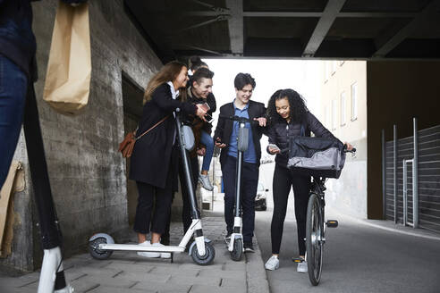 Fröhliche männliche und weibliche Teenager-Freunde mit Elektro-Scootern, die unter einer Brücke auf ihr Handy schauen - MASF12865