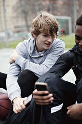 Jugendlicher schaut auf das Handy seines Freundes, während er nach dem Basketballtraining auf dem Gehweg sitzt - MASF12906