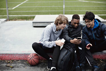 Männliche Freunde benutzen Mobiltelefone, während sie nach dem Basketballtraining im Winter auf dem Bürgersteig sitzen - MASF12907