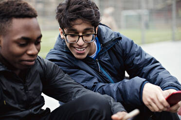 Lächelnder junger Mann, der auf das Mobiltelefon seines Freundes schaut, auf einem Spielfeld in der Stadt im Winter - MASF12909