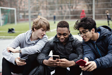 Lächelnde Freunde, die auf das Handy eines Teenagers schauen, während sie auf einem Fußballfeld sitzen - MASF12912