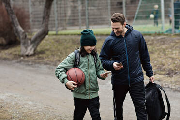 Glücklicher Vater und Sohn benutzen Mobiltelefone beim Spaziergang auf der Straße nach dem Basketballtraining im Winter - MASF12915