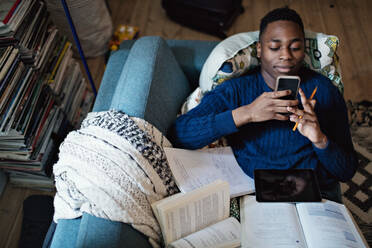 High angle view of teenage boy using mobile phone while lying on sofa with books at home - MASF12930