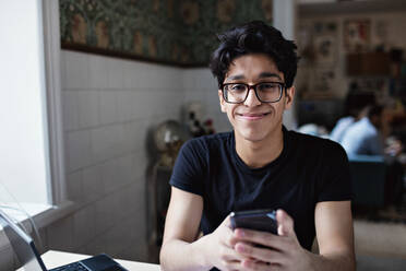 Porträt eines lächelnden jungen Mannes, der soziale Medien auf seinem Mobiltelefon nutzt, während er zu Hause lernt - MASF12944