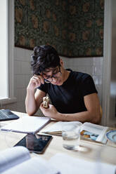 Studious young man holding sandwich while reading book on table at home - MASF12947