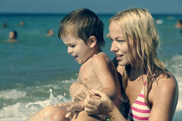 Eine Welle spritzt auf eine Mutter, die ihren Sohn am Strand hält - BLEF09692