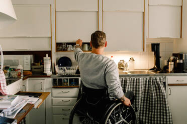 Rear view of disabled man opening cabinet in kitchen at home - MASF12996