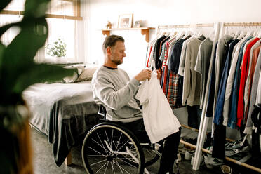 Side view of disabled mature man holding white t-shirt while sitting on wheelchair by clothes rack at home - MASF13002