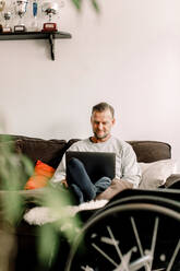 Disabled mature man using laptop while sitting on sofa at home - MASF13005