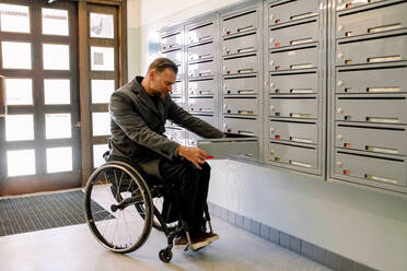 Behinderter reifer Mann in voller Länge beim Öffnen eines Briefkastens im Umkleideraum, während er im Rollstuhl sitzt - MASF13006